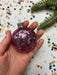 A hand holding a pink glitter ornament against a background of colorful confetti and a pine tree branch.