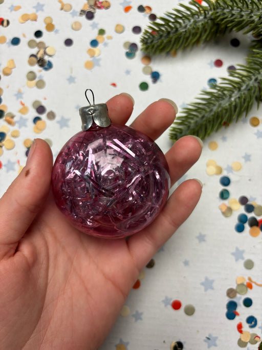 A hand holding a pink glitter ornament against a background of colorful confetti and a pine tree branch.