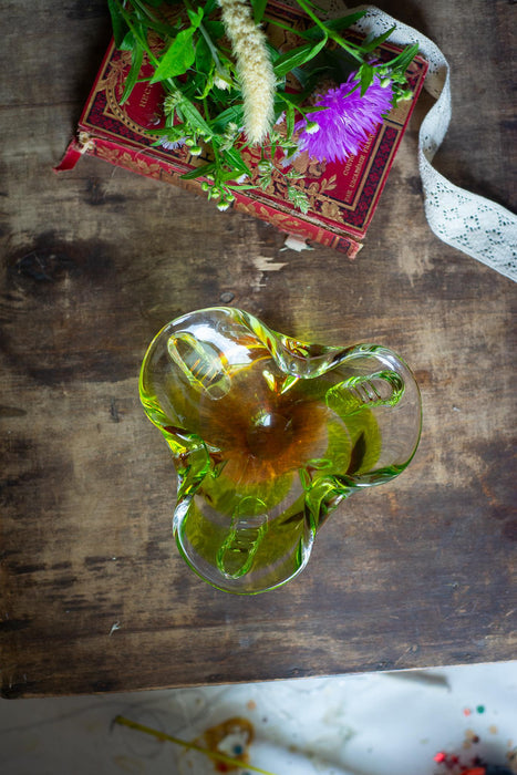 a green glass flower vase with a purple flower, placed on a wooden surface next to a red book with a white ribbon.