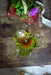 a green glass flower vase with a purple flower, placed on a wooden surface next to a red book with a white ribbon.