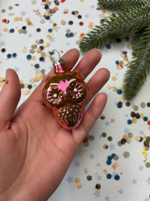 A hand holding a small, shiny ornament with a pink and green design, surrounded by colorful confetti on a white background.