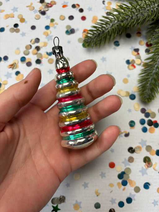 A hand holding a colorful, multi-colored ornament against a background of confetti and a pine branch.