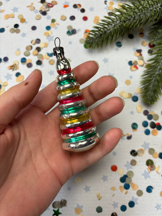 A hand holding a colorful, multi-colored ornament against a background of confetti and a pine branch.