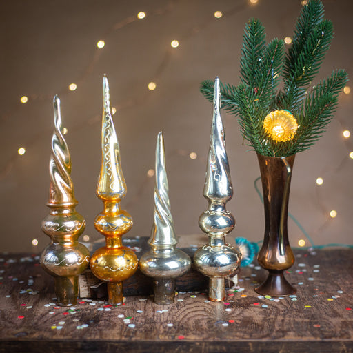 a collection of ornate Christmas tree ornaments, including golden and silver spires, placed on a wooden surface. A vase with a green plant and a single yellow flower is also present, creating a festive and decorative scene.