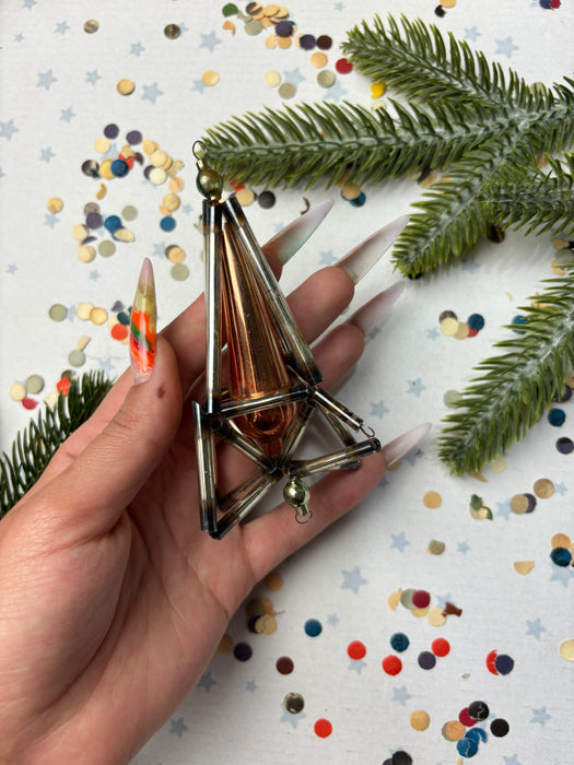 A hand holding a metallic ornament with a pointed top, surrounded by Christmas tree branches and confetti on a white background.