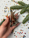 A hand holding a metallic ornament with a pointed top, surrounded by Christmas tree branches and confetti on a white background.