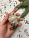 A hand holding a small, white owl ornament with red accents against a background of colorful confetti and a pine tree branch.