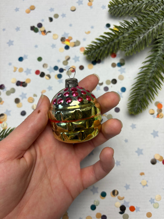 A hand holding a small, colorful ornament with a shiny, metallic appearance, surrounded by scattered confetti and a sprig of greenery.